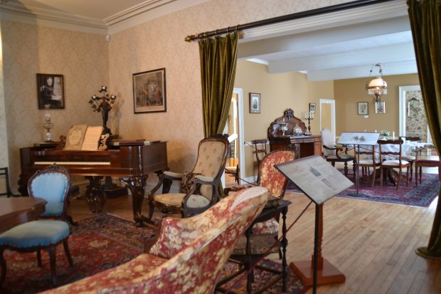 Photo couleur. Salon aménagé dans le style victorien, dans le coin un piano table, en arrière-plan une salle à manger, à l'avant-plan un panneau d'interprétation.