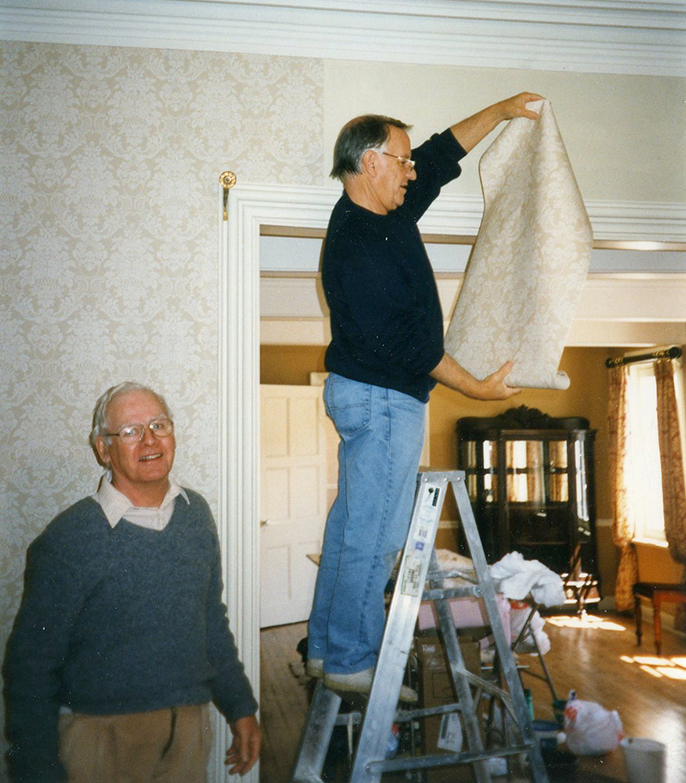 Photo couleur. Deux hommes, dont un debout sur un escabeau, posent de la tapisserie dans le salon d’apparat.