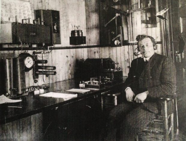 Un homme en complet est assis face à l’appareil-photo, à côté d'un bureau sur lequel sont posés du matériel de télégraphie sans fil et des piles de papier soigneusement ordonnées. Il porte un casque d'écoute et fume la pipe.