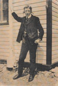 Un jeune homme portant un complet et un gilet se tient devant un bâtiment de bardage à clin, son bras droit levé tenant un mât porte-drapeau. Il porte des écouteurs et regarde directement vers l'appareil photo.