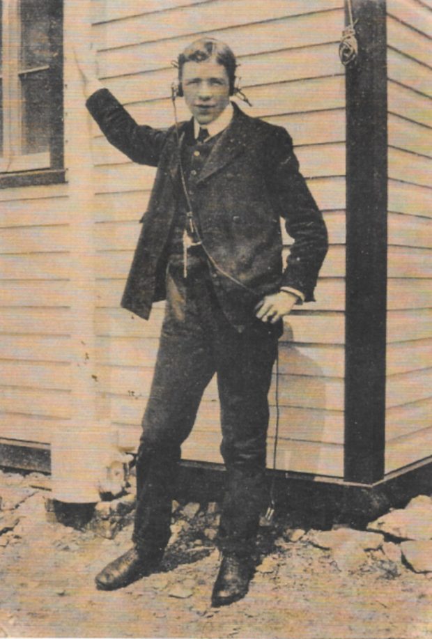 Un jeune homme portant un complet et un gilet se tient devant un bâtiment de bardage à clin, son bras droit levé tenant un mât porte-drapeau. Il porte des écouteurs et regarde directement vers l'appareil photo.