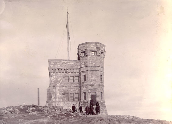Un groupe de personnes se tient devant un bâtiment en pierre de deux étages situé sur une colline rocheuse aride. Le bâtiment comporte une tourelle plus haute sur la droite et un mât sur le toit à gauche.
