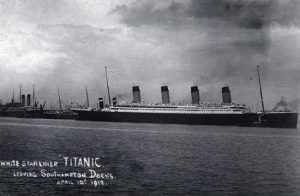 Un grand navire à quatre cheminées quitte un port par la droite, laissant derrière lui des navires plus petits. La photographie porte l'inscription suivante : « Paquebot de la White Star Line 'Titanic'. Quittant les quais de Southampton, le 10 avril 1912. » (Traduction)