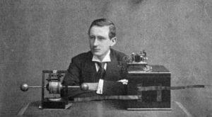 Un jeune homme est assis, les bras croisés appuyés sur une table. Du matériel radio est posé sur la table devant lui.