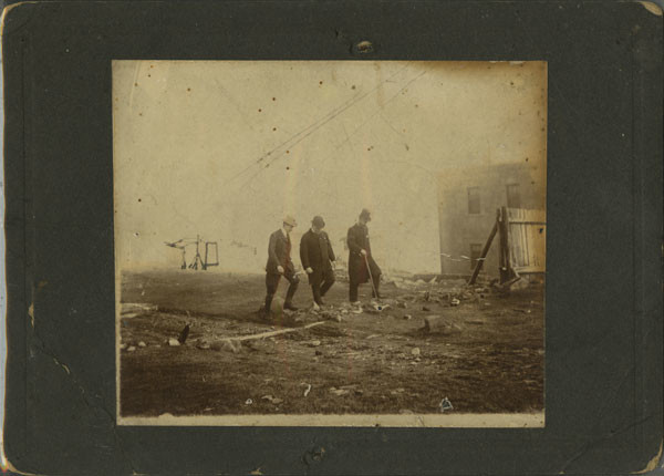 Trois hommes portant complets et chapeaux marchent ensemble sur une colline d'herbe rase et de rochers.