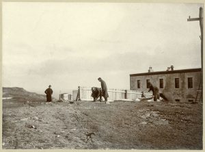 Quatre hommes installent un grand cerf-volant sur le sol. Derrière eux se trouvent une clôture en bois et un vieux bâtiment en béton. Au loin, on aperçoit des collines et l'océan.