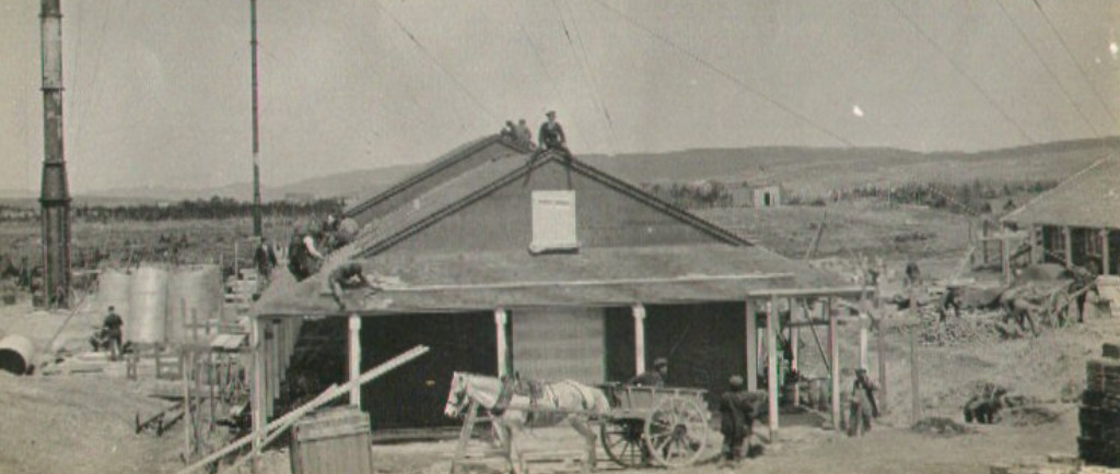 Bâtiment en bois de plain-pied avec un cheval et une charrette devant. Des ouvriers travaillent autour du bâtiment et sur le toit.