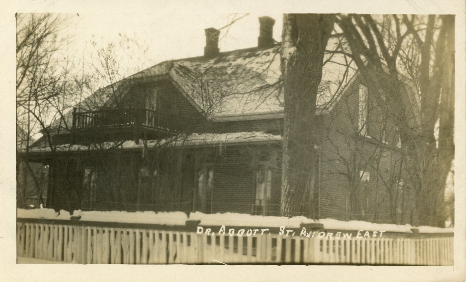 Photographie d’Elmbank, la résidence de Maude Abbott, en hiver, sépia. Il s’agit d’une maison en briques de deux étages avec un toit à deux versants, deux cheminées et une galerie à chaque étage. On aperçoit une clôture en bois blanche à l’avant de la maison, qui est entourée d’arbres sans feuille. 