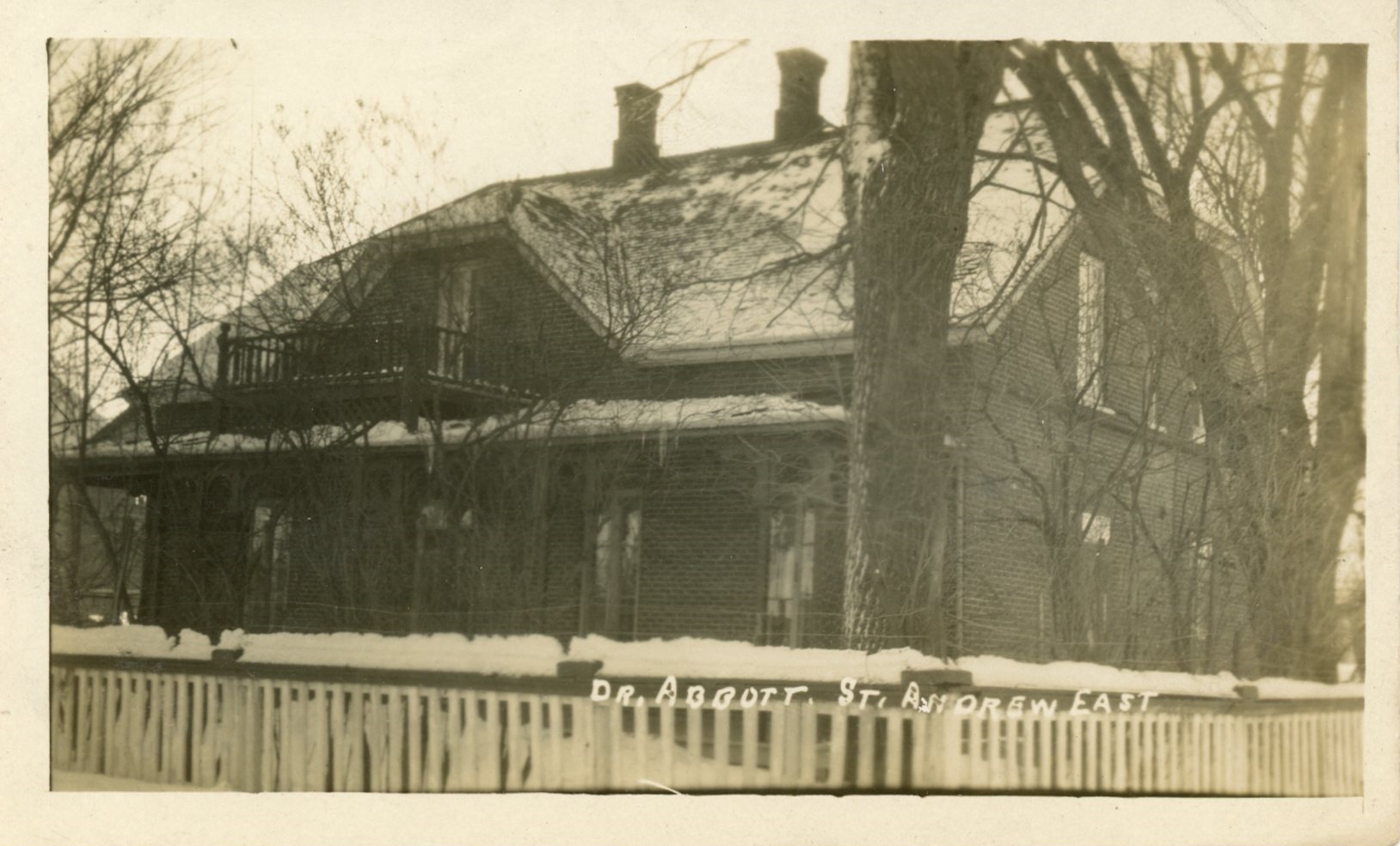 Photographie d’Elmbank, la résidence de Maude Abbott, en hiver, sépia. Il s’agit d’une maison en briques de deux étages avec un toit à deux versants, deux cheminées et une galerie à chaque étage. On aperçoit une clôture en bois blanche à l’avant de la maison, qui est entourée d’arbres sans feuille.