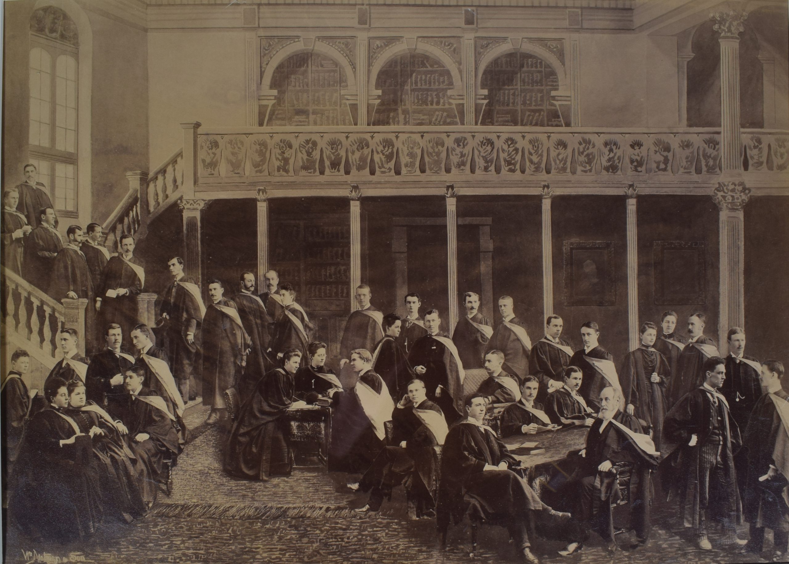 Photographie d’une classe de jeunes adultes, hommes et quelques femmes, sépia. On compte 35 étudiants dispersés dans une salle. Ils portent tous une toge de graduation noire avec une écharpe blanche aux rebords foncés sur l’épaule gauche. Les hommes ont les cheveux courts et les femmes ont les cheveux attachés à l’arrière de la tête. Ils sont dans une salle avec un grand tapis au sol, devant un balcon et des escaliers à colonnes et de grandes bibliothèques. 