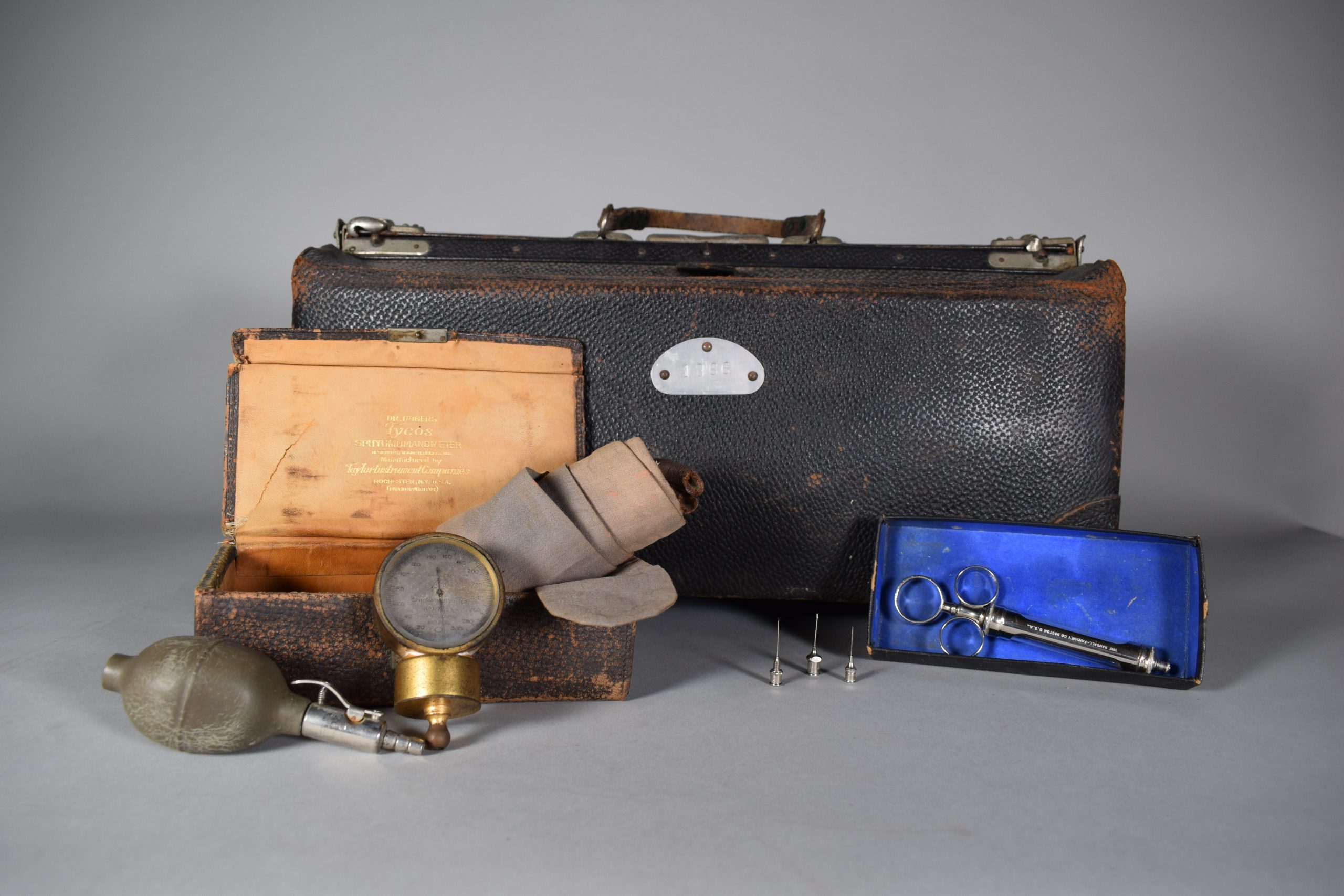 Photographie en couleur d’une trousse médicale et d’instruments médicaux. À l’arrière, la grande trousse rectangulaire en cuir noir, avec poignée au centre et serrures des deux côtés. La trousse est légèrement abimée aux coins.   En avant de la trousse, à gauche, une plus petite trousse, elle aussi en cuir noir. Elle est ouverte, l’intérieur est beige. À l’intérieur de cette trousse, dépassant du côté droit, un bandage en coton beige-gris enroulé autour de supports en métal rouillé. Une poire de tensiomètre et un sphygmomanomètre y sont appuyés. À droite, une ancienne seringue est placée dans une boîte en carton et on peut voir, à sa gauche, trois aiguilles. Le sphygmomanomètre doré, appuyé sur la trousse ouverte.