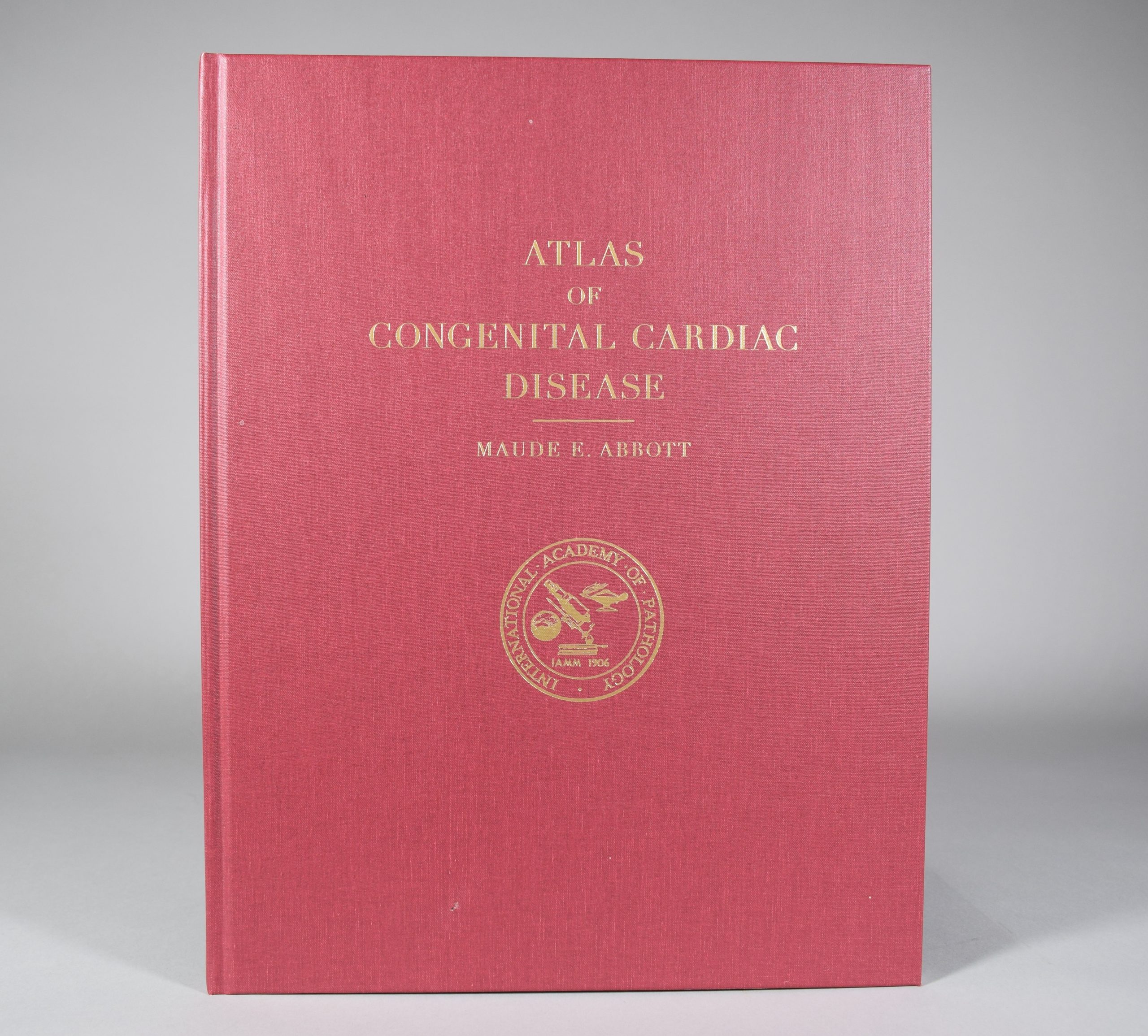 Photographie en couleur de l’Atlas de Maude Abbott. Il est rouge avec l’inscription « Atlas of Congenital Cardiac Disease – Maude E. Abbott » en lettres dorées. Sous le titre, le sceau doré de l’International Academy of Pathology : le nom de l’académie entoure des images d’un globe terrestre, d’un microscope et une lampe merveilleuse, au-dessus de l’inscription « IAMM 1906 ». 