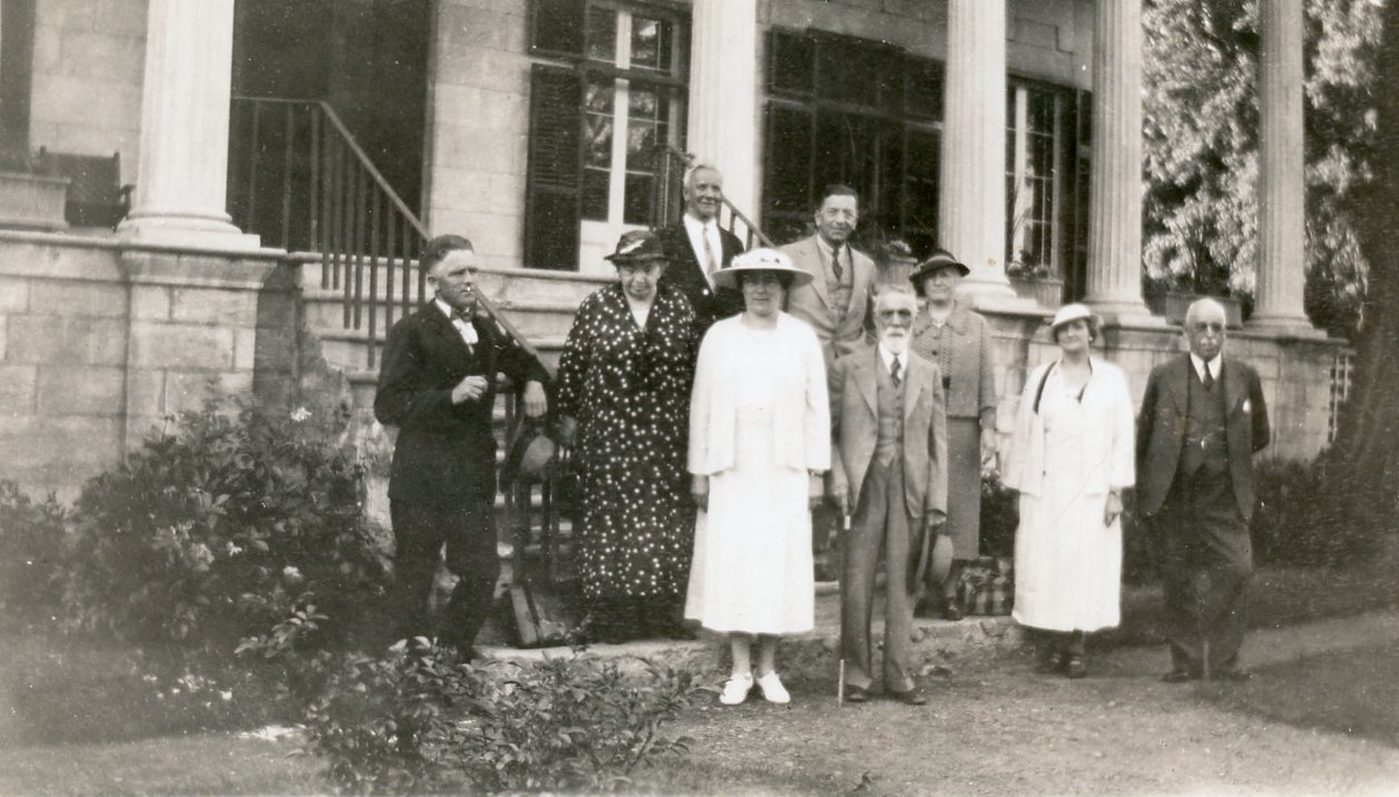 Photographie en noir et blanc des neuf membres fondateurs de la Société historique du comté d’Argenteuil. Ils sont debout devant et dans les escaliers d’une grande maison en pierre avec colonnes et volets foncés. Maude Abbott est la deuxième à partir de la gauche, portant une robe noire à pois blancs et un chapeau foncé. Les trois autres femmes sont vêtues de robes et chapeaux chics et les cinq hommes portent des complets.