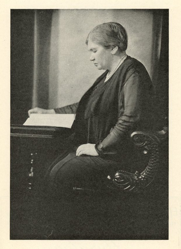 Photographie de Maude Abbott, âgée, noir et blanc. Elle est vue de profil, l’air concentré, lisant un livre à un bureau en bois et assise sur un siège en bois sculpté. Elle porte des vêtements foncés, un collier de perles et ses cheveux sont attachés derrière sa tête.