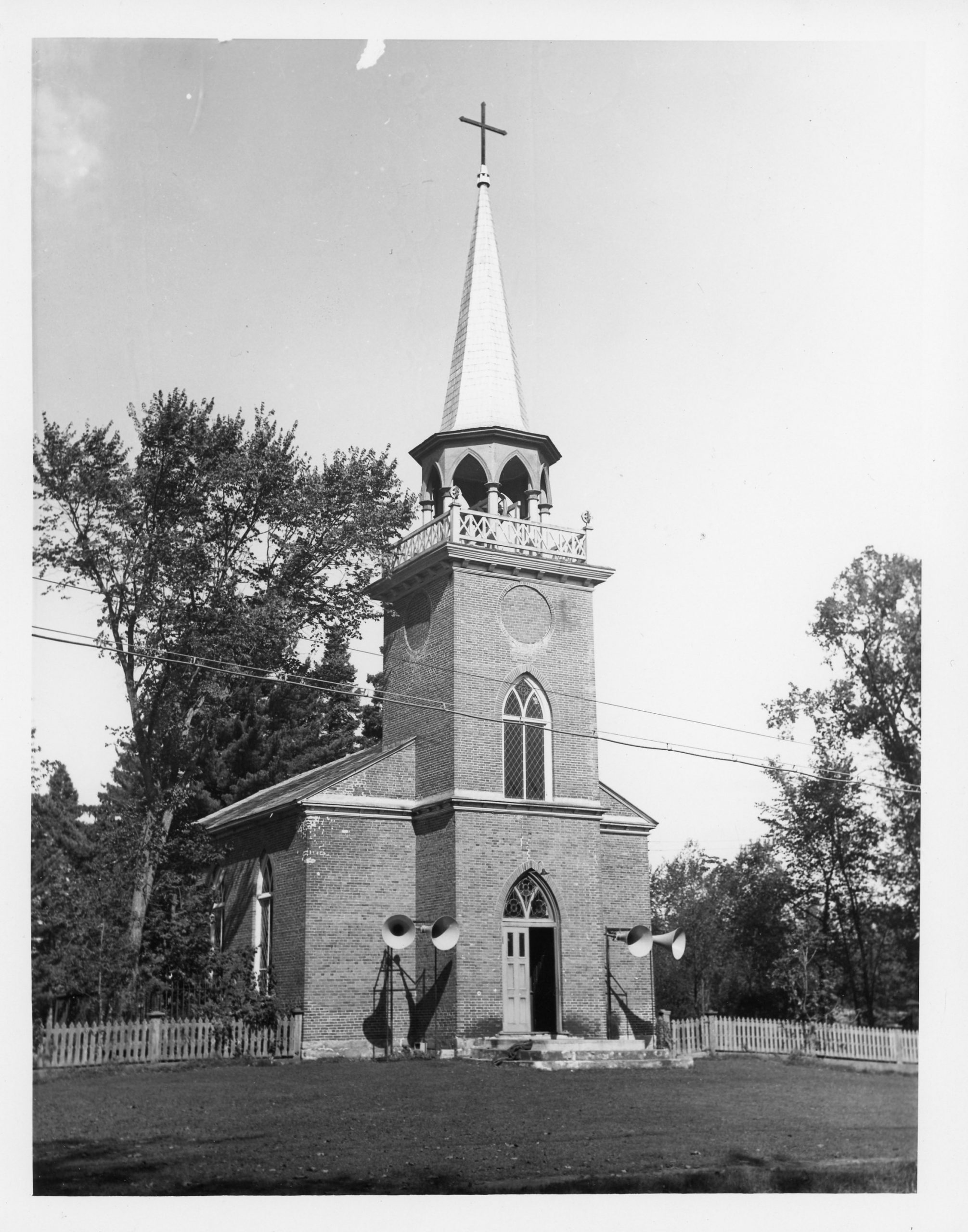 Photographie en noir et blanc de l’église Christ Church. L’église est en brique, la porte double, au centre, est ouverte d’un côté et surmontée d’une fenêtre en ogive. Une autre fenêtre de la même forme se trouve dans la tour du clocher. Les ouvertures du clocher sont en forme d’ogive et il est surmonté d’une longue flèche au bout de laquelle se trouve une croix noire. Deux haut-parleurs sont installés de chaque côté de la porte et beaucoup d’arbres se trouvent à l’arrière de l’église.