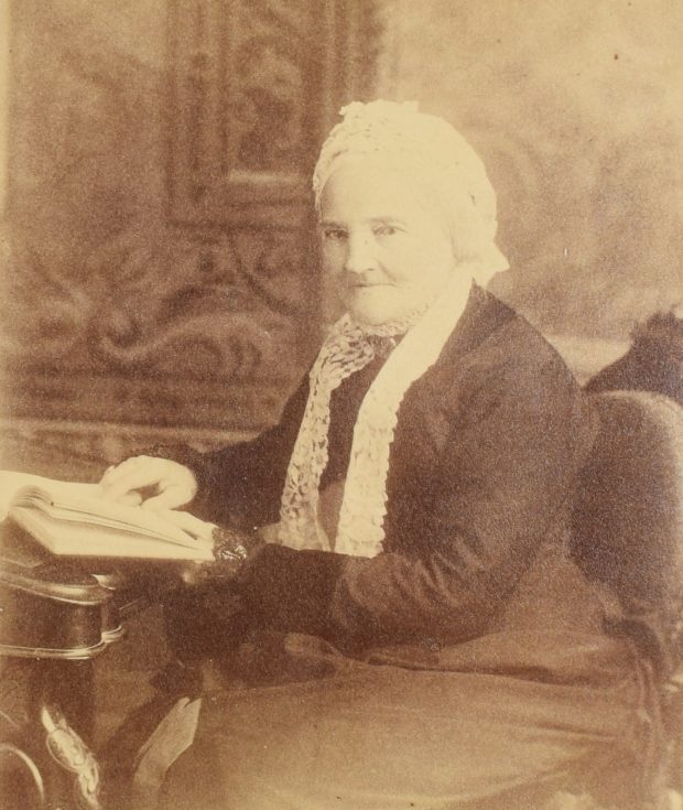 Photographie sépia de Frances Mary Smith âgée, en 1880, assise, des genoux à la tête. Elle regarde l’objectif, la tête légèrement tournée, l’air serein. Elle est appuyée sur un bureau, la main gauche sur un livre ouvert. Elle porte une robe noire et un bonnet en dentelle blanc. Ses cheveux blancs sont attachés à l’arrière de sa tête, sous le bonnet.
