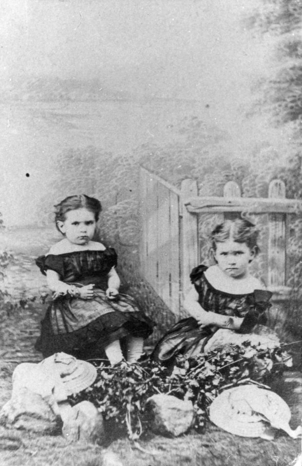 Photographie en noir et blanc de Maude et sa sœur Alice enfants. Elles sont assises au bord d’une clôture et regardent l’objectif avec un air sérieux. Elles portent des robes aux épaules dénudées et manches bouffantes foncées. Maude, à gauche, a les cheveux foncés, attachés à l’arrière de sa tête et porte une fleur à son bras droit. Alice, à droite, a les cheveux pâles, également attachés à l’arrière de sa tête. Elles sont assises derrière un arrangement de fleurs, deux chapeaux et trois roches sur la pelouse.