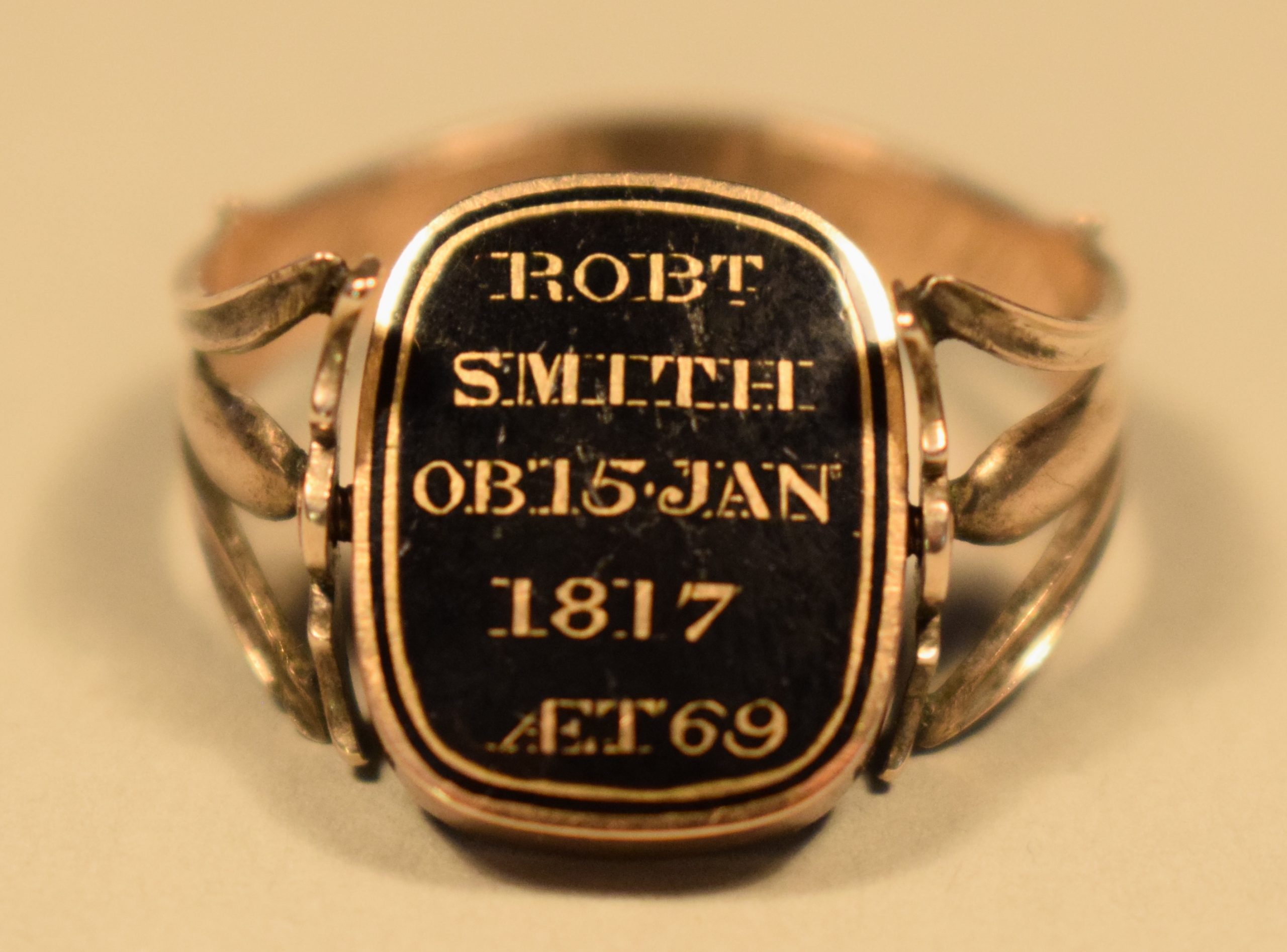 Photographie en couleur de la face d’une bague en or rose. La bague est de couleur or et l’élément décoratif est un détail au fond noir et aux lettres et bordures or avec l’inscription « ROBT SMITH OB 15 JAN 1817 AET 69 ». Le fond derrière et sous la bague est beige.