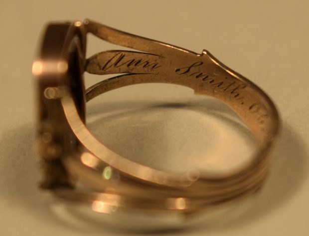 Photographie en couleur de l’intérieur du côté gauche d’une bague en or rose. La bague est de couleur or et on y voit l’inscription « Ann Smith » (voir la photographie de l’intérieur arrière de la bague pour la suite) gravée. Le fond derrière et sous la bague est beige.