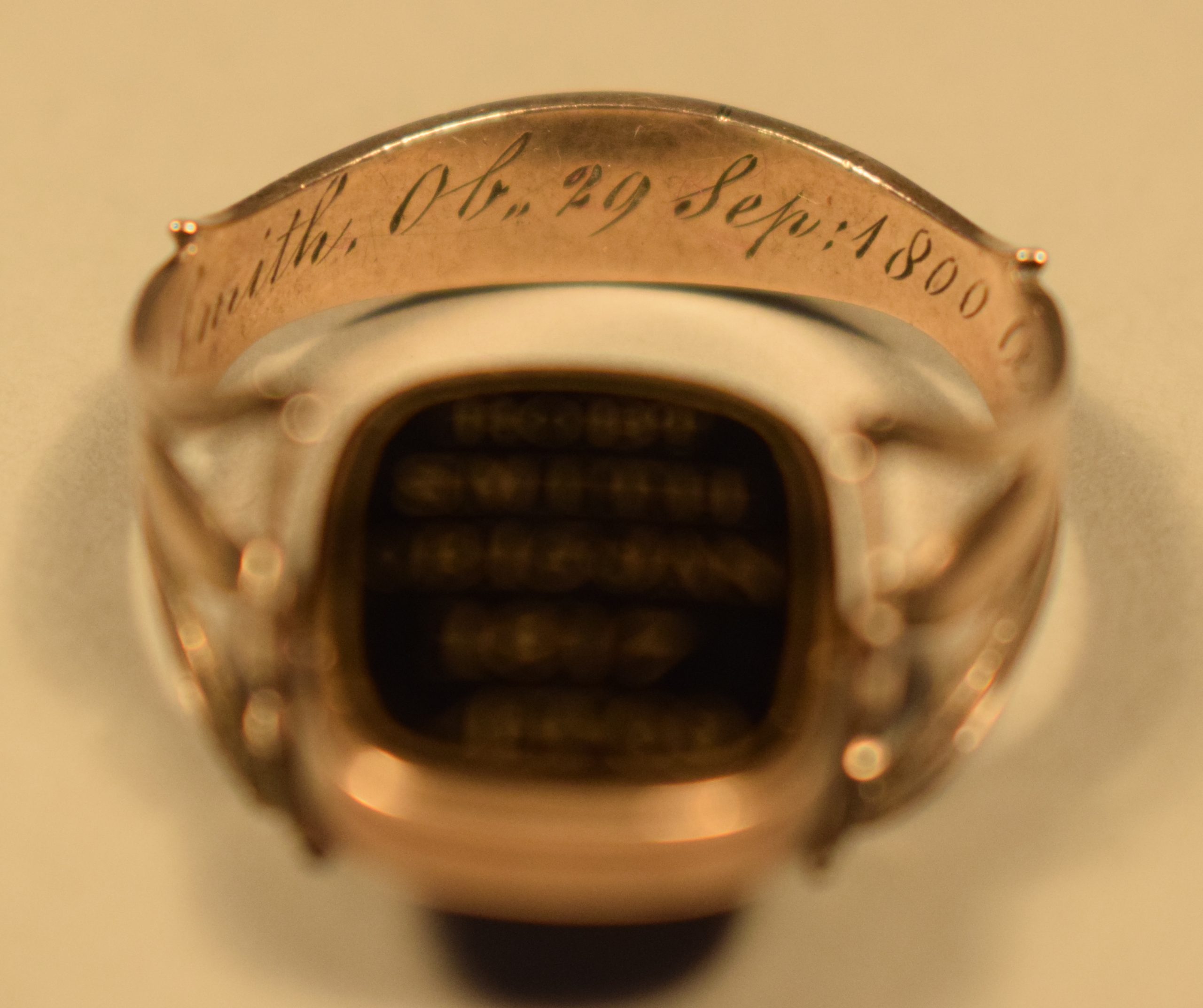 Photographie en couleur de l’intérieur de l’arrière d’une bague en or rose. La bague est de couleur or et on peut y lire l’inscription « Smith Ob,, 29 sep : 1800 » (voir la photographie de l’intérieur du côté droit pour la suite) gravée. Le fond derrière et sous la bague est beige.