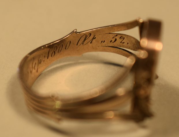 Photographie en couleur de l’intérieur du côté droit d’une bague en or rose. La bague est de couleur or et on y voit l’inscription « Sep : 1800 aet ,, 52 » gravée. Le fond derrière et sous la bague est beige.
