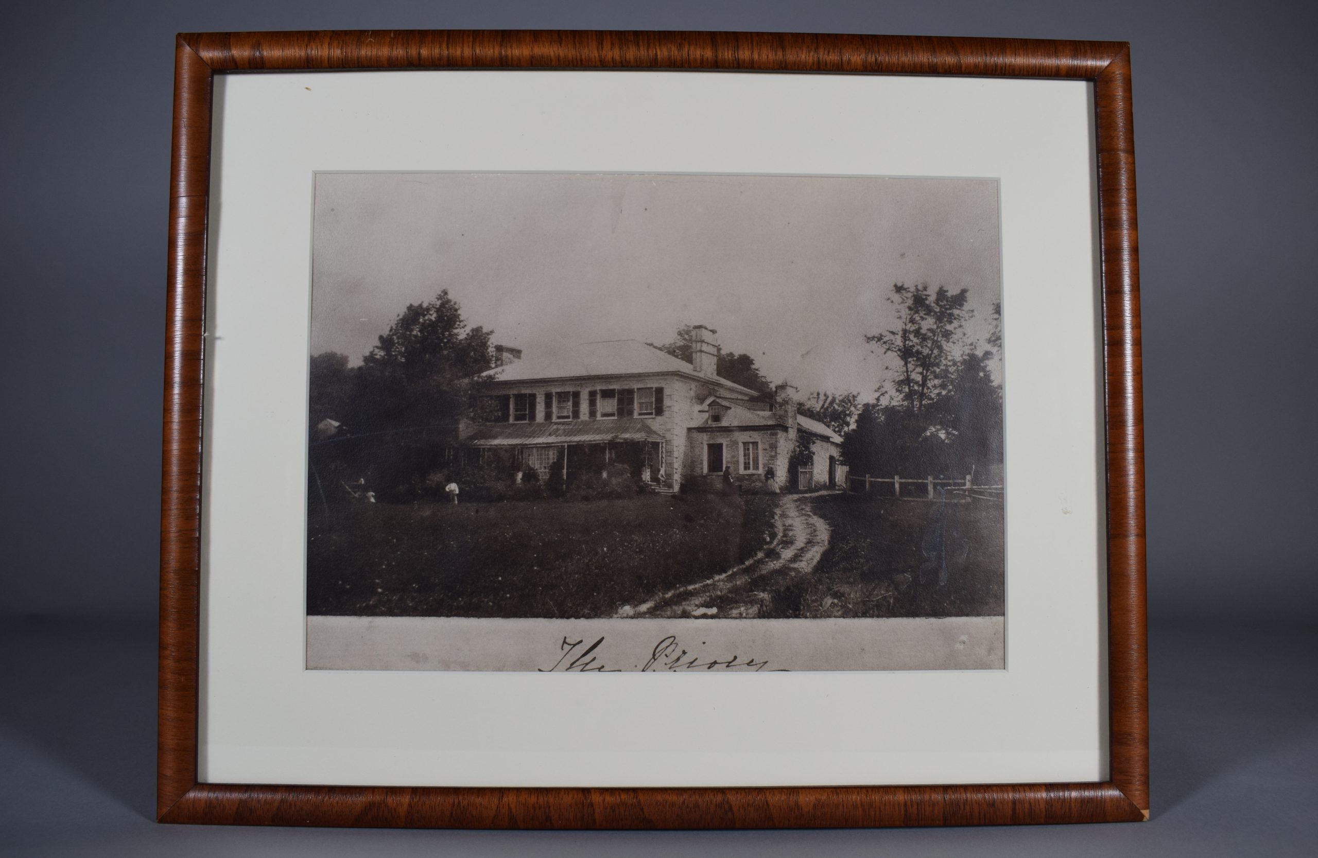 Photographie en noir et blanc d’une maison à deux étages, entourée d’arbres, l’été. Elle est en briques grises avec une grande galerie. L’étage a cinq fenêtres à volets foncés et on aperçoit deux cheminés au toit. Une section est adjacente à la maison, un peu reculée et son toit s’arrêtant aux trois quarts de l’étage de la maison. Deux femmes sont debout devant la partie adjacente de la maison, un homme est assis dans les escaliers et un autre est dans la cour devant la maison. Sous la photo, « The Priory » est inscrit à l’encre noire. La photographie est placée dans un mince cadre en bois simple, avec un passe-partout blanc.