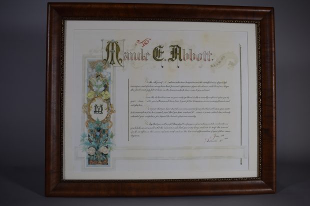Certificat de reconnaissance en couleur. En haut, « Maude E. Abbott » dont les initiales sont en lettrines et dans une bannière à droite « B.A., C.M., M.D. ». Du côté gauche, une banderole avec des fleurs dans un encadré noir entoure un cercle doré avec les lettres « MD » superposées. 