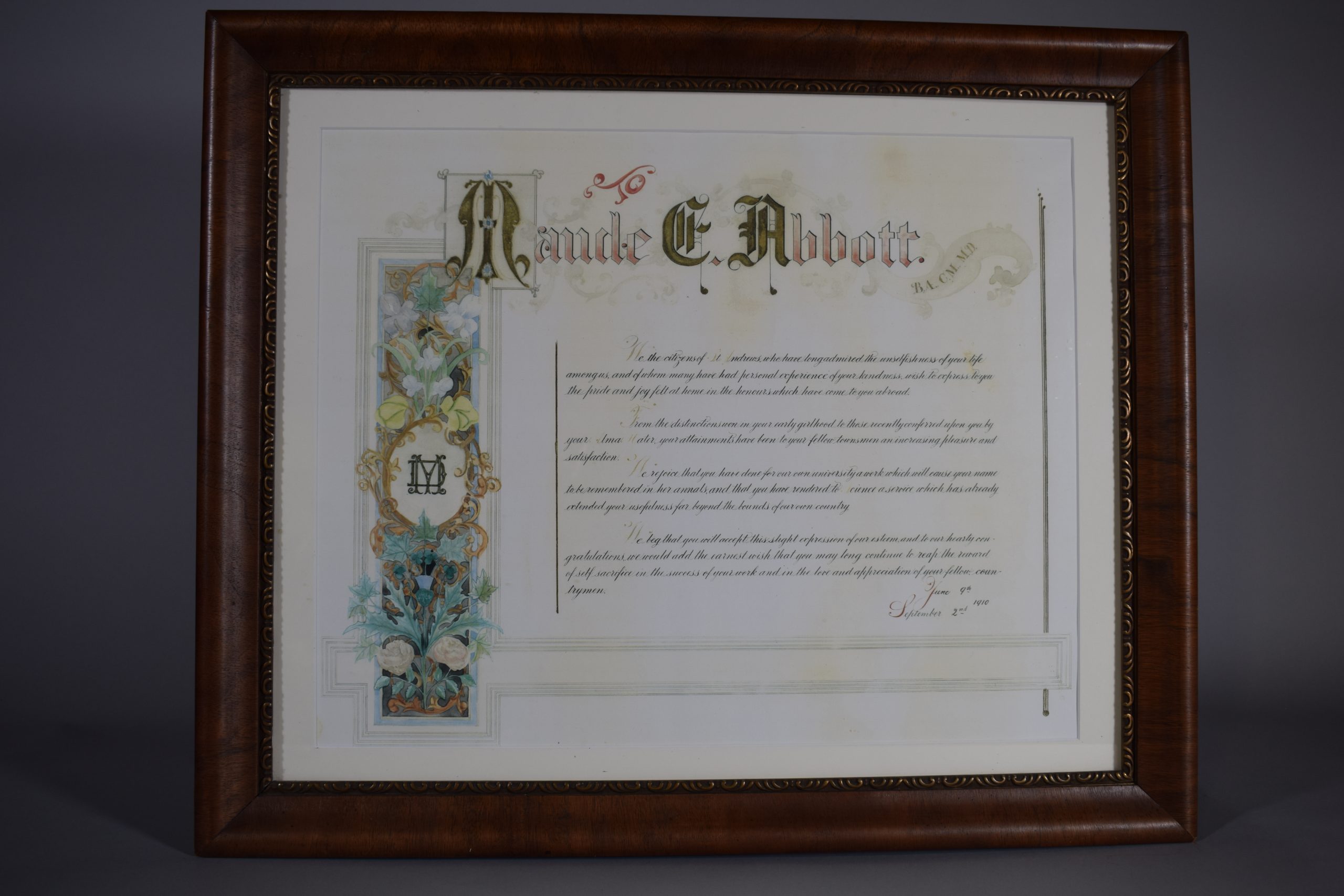 Certificat de reconnaissance en couleur. En haut, « Maude E. Abbott » dont les initiales sont en lettrines et dans une bannière à droite « B.A., C.M., M.D. ». Du côté gauche, une banderole avec des fleurs dans un encadré noir entoure un cercle doré avec les lettres « MD » superposées. 