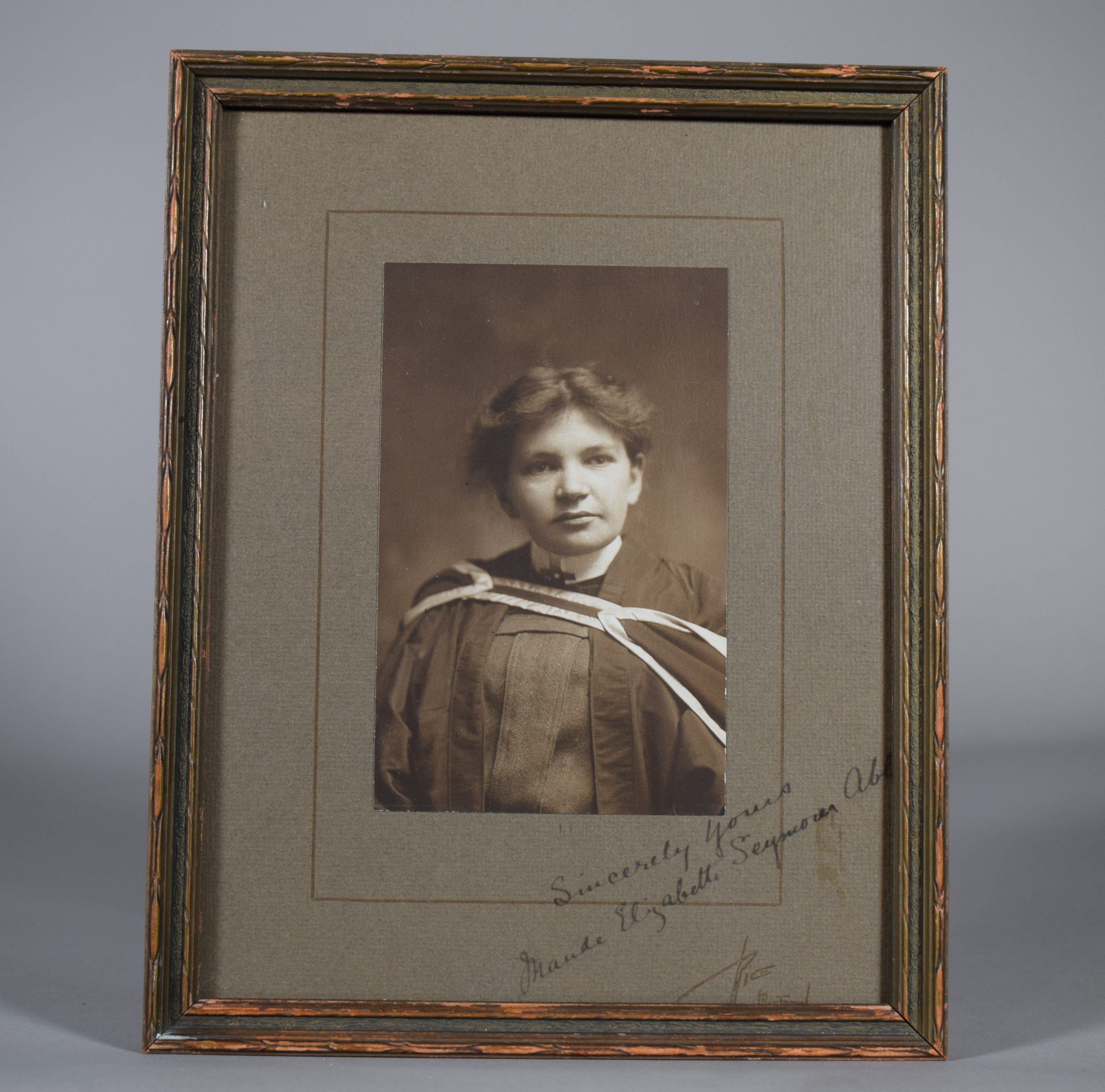 Photographie en noir et blanc de Maude Abbott, jeune adulte, de la taille à la tête. Elle porte un habit et une toge de graduation. Ses cheveux foncés sont attachés à l’arrière de sa tête et sa tête est légèrement tournée vers la gauche. Dans le coin inférieur droit, une signature à l’encre : « Sincerely Yours. Maude Elizabeth Seymour Abbott ». Un peu plus bas, la signature de l’imprimeur, « Rice. Montreal ». 