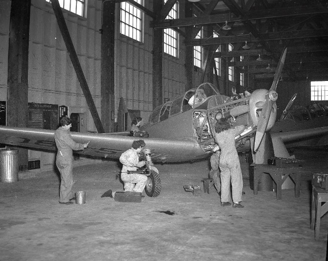 Une photographie en noir et blanc de cinq mécaniciennes qui réparent un avion dans un hangar. Une personne est assise dans l’avion sur le siège du pilote.   