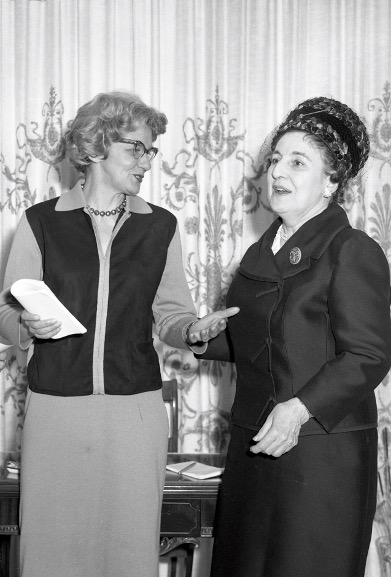 Photographie en noir et blanc d’Adrienne Choquette et Claire Roy qui discutent. Les deux femmes sont debout devant un bureau. 