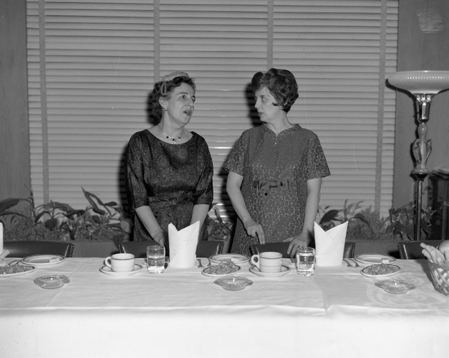 Photographie en noir et blanc de deux femmes debout derrière une table. Claire Roy (gauche) discute avec l’autre femme. De la vaisselle repose sur la table devant elles et une lampe est visible à droite de la photographie. 