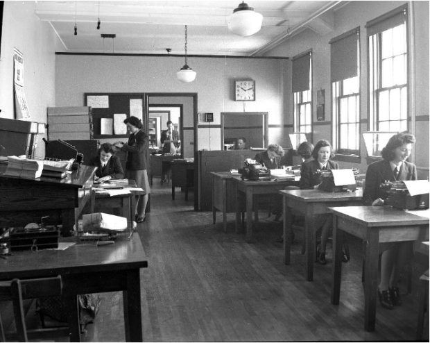 Photographie en noir et blanc de femmes participant à l’effort de guerre. À droite de la photographie, des femmes assises à des tables rédigent des lettres avec une dactylo. À gauche, une femme est assise à un bureau rempli de paperasse et une femme cherche dans un classeur. Dans le fond de la pièce, une porte ouverte permet de voir un homme debout devant un bureau dans une pièce adjacente.  