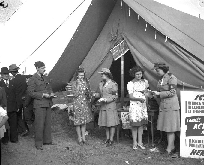 Photographie en noir et blanc de quatre femmes dont deux habillées en uniforme discutent devant une tente du bureau de recrutement de l’armée. À gauche de la photographie, un homme en uniforme est debout devant trois personnes.    