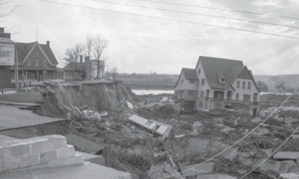 Photographie en noir et blanc d’un glissement de terrain. À gauche de la photographie se trouve les maisons ayant survécu au désastre. À droite, une maison touchée par le glissement de terrain repose plus bas parmi les ruines.  