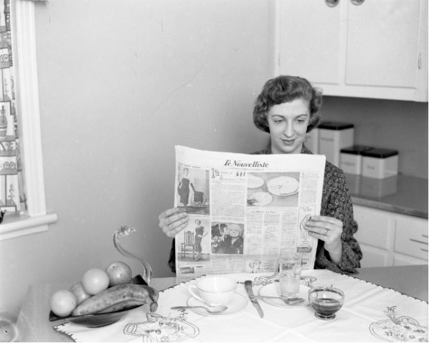 Photographie en noir et blanc d’une femme assise à la table d’une cuisine qui lit le journal Le Nouvelliste. Une tasse, deux verres, deux cuillères, un couteau et un bol en forme de cygne contenant des fruits reposent sur la table devant elle.  