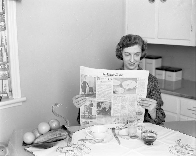 Black and white photograph of a woman sitting at the kitchen table, reading Le Nouvelliste. A cup, two glasses, two spoons, a knife and a swan-shaped bowl with fruit are on the table in front of her.