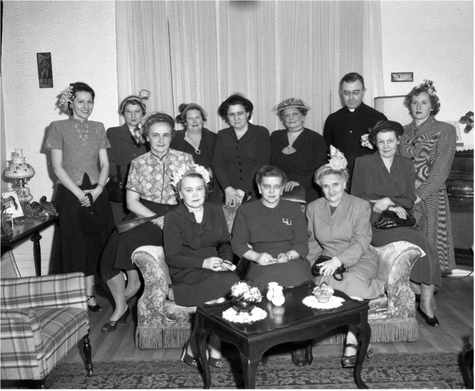 Photographie de membres de la ligue catholique féminine de la Mauricie avec un membre du clergé. Trois femmes sont assises sur un divan alors que huit femmes et un membre du clergé sont derrières elles. Une table basse est devant eux et un siège se trouve à leur droite.  