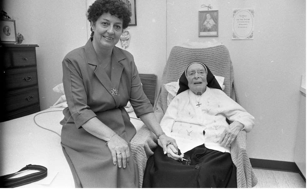 Black and white photograph of a nun sitting on a chair at the Ursuline Monastery. She is holding the hand of Michelle Roy, who is sitting on a bed to the nun’s right.