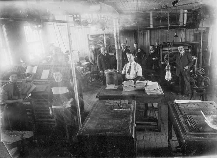 Blurry black and white photograph showing the newsroom at La Semaine. In the back, six men and one woman are standing near a printing press. In the centre, a man is standing behind three tables covered with newspapers. On the left, a woman and another person are leaning against a desk.