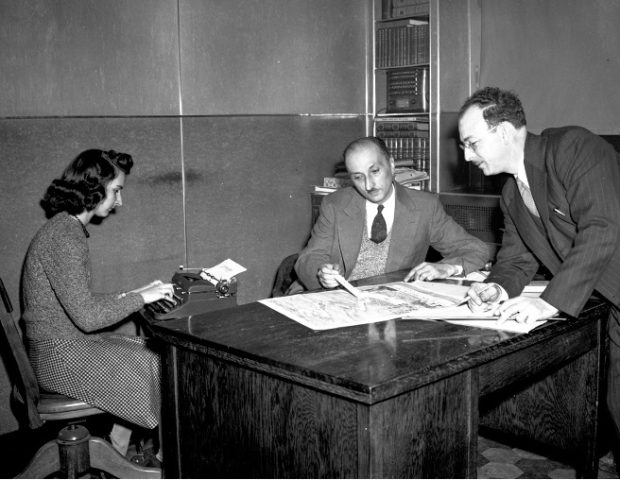 Photographie en noir et blanc d’une femme qui dactylographie la conversation entre Émile Jean, directeur-gérant et Jos Lefebvre, directeur du service de publicité locale. Émile Jean, assis à un bureau, pointe sur une carte alors que Jos Lefebvre, debout à droite du bureau regarde la carte.  