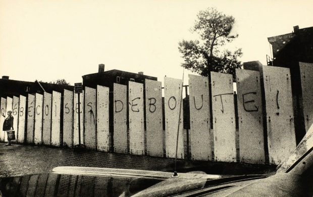 Photographie en noir et blanc qui montre des panneaux qui ont été peints avec l’écriture ‘’Québécoises Debouute!’’. Une femme se tient debout à côté des panneaux.