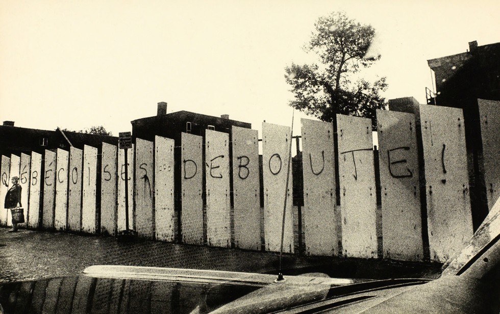 Photographie en noir et blanc qui montre des panneaux qui ont été peints avec l’écriture ‘’Québécoises Debouute!’’. Une femme se tient debout à côté des panneaux.