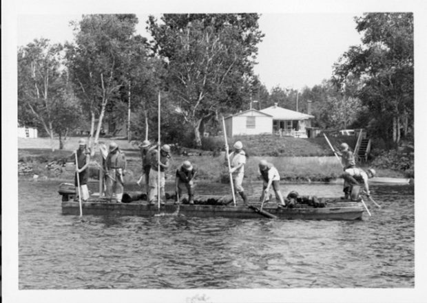 Photographie en noir et blanc qui montrent des gens faire la drave sur la rivière Saint-Maurice en 1987. Neuf personnes sont sur la photo et se tiennent sur un ponton avec leurs perches à la main.