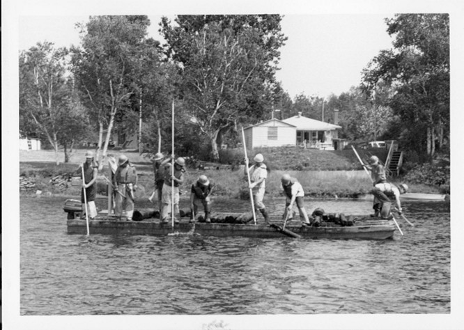 Photographie en noir et blanc qui montrent des gens faire la drave sur la rivière Saint-Maurice en 1987. Neuf personnes sont sur la photo et se tiennent sur un ponton avec leurs perches à la main.