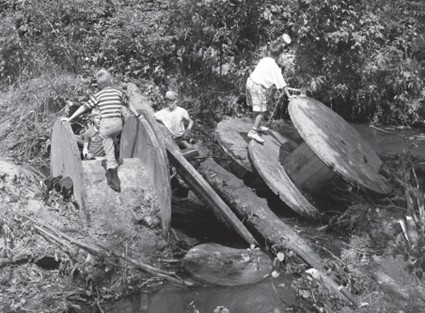 Photographie en noir et blanc qui montre des enfants jouant sur des pièces d’équipement laissé par une ancienne compagnie lors d’une opération de nettoyage des berges de la Petite rivière Champlain en août 1989.