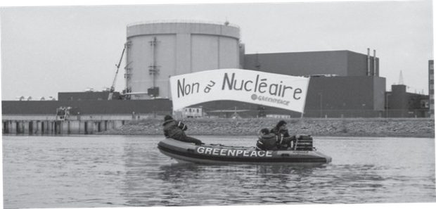 Photographie en noir et blanc montrant trois militants du regroupement écologiste Greenpeace dans un zodiac sur le fleuve Saint-Laurent devant la centrale Gentilly-2. Les militants tiennent dans leurs mains une banderole avec l’écriture ‘’Non au nucléaire’’.
