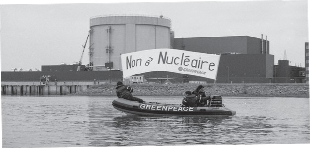 Photographie en noir et blanc montrant trois militants du regroupement écologiste Greenpeace dans un zodiac sur le fleuve Saint-Laurent devant la centrale Gentilly-2. Les militants tiennent dans leurs mains une banderole avec l’écriture ‘’Non au nucléaire’’.