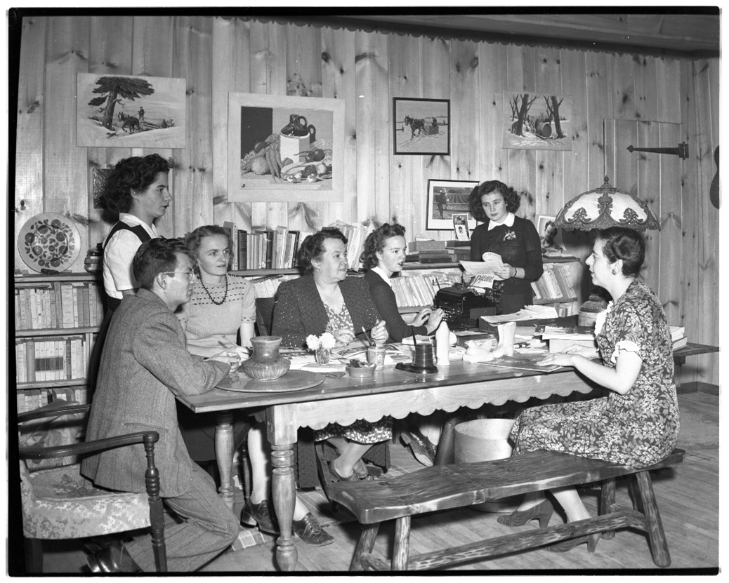 Photographie en noir et blanc qui montre six femmes et un homme assis à une table à l’intérieur d’une maison en 1946.
