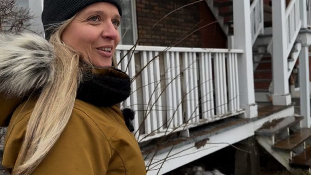 Photographie en couleur de Paule Vermot-Desroches habillée d’un manteau d’hiver jaune et d’une tuque marchant devant le balcon d’un immeuble à appartements. 